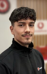 Ein junger Mann mit dunklen Locken und einem leichten Lächeln trägt eine schwarze Jacke.